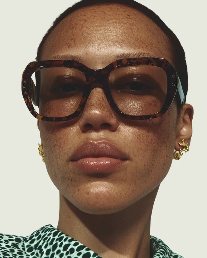 Close-up portrait of a woman with freckles wearing large tortoiseshell glasses, gold earrings, and a green patterned shirt.